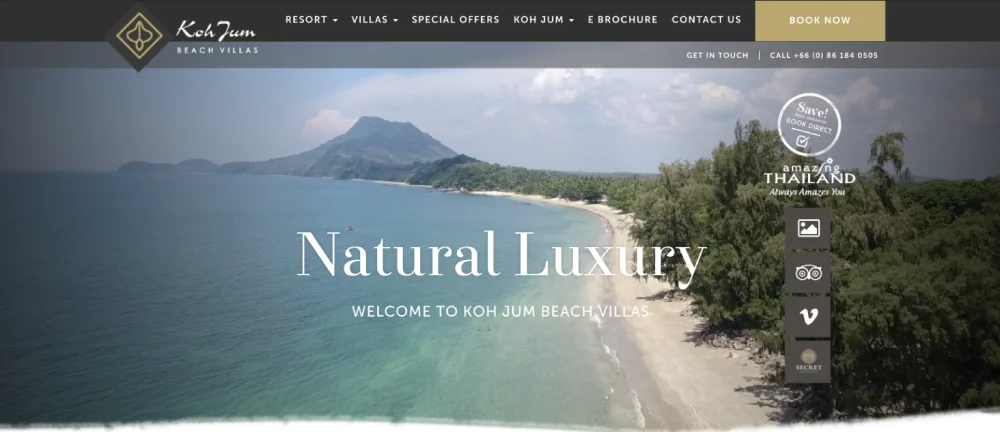 Aerial view of Koh Jum Beach Villas with "Natural Luxury" gracing the image. Showcasing lush greenery, a sandy beach, and a distant mountain, this epitomizes the best hotel website design. Contact and booking options are conveniently displayed at the top.