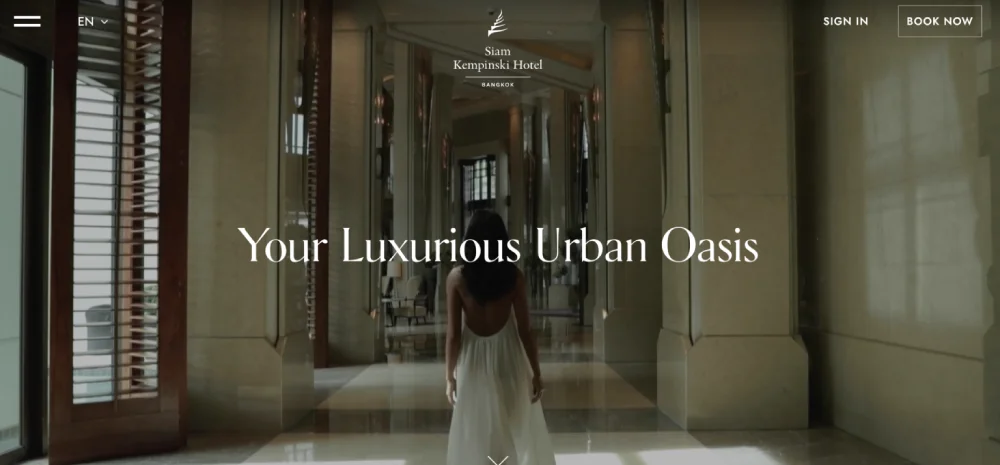 A woman in a white dress walks through a grand hallway with large windows, epitomizing elegance. Text reads: "Your Luxurious Urban Oasis – showcasing the best hotel website design.